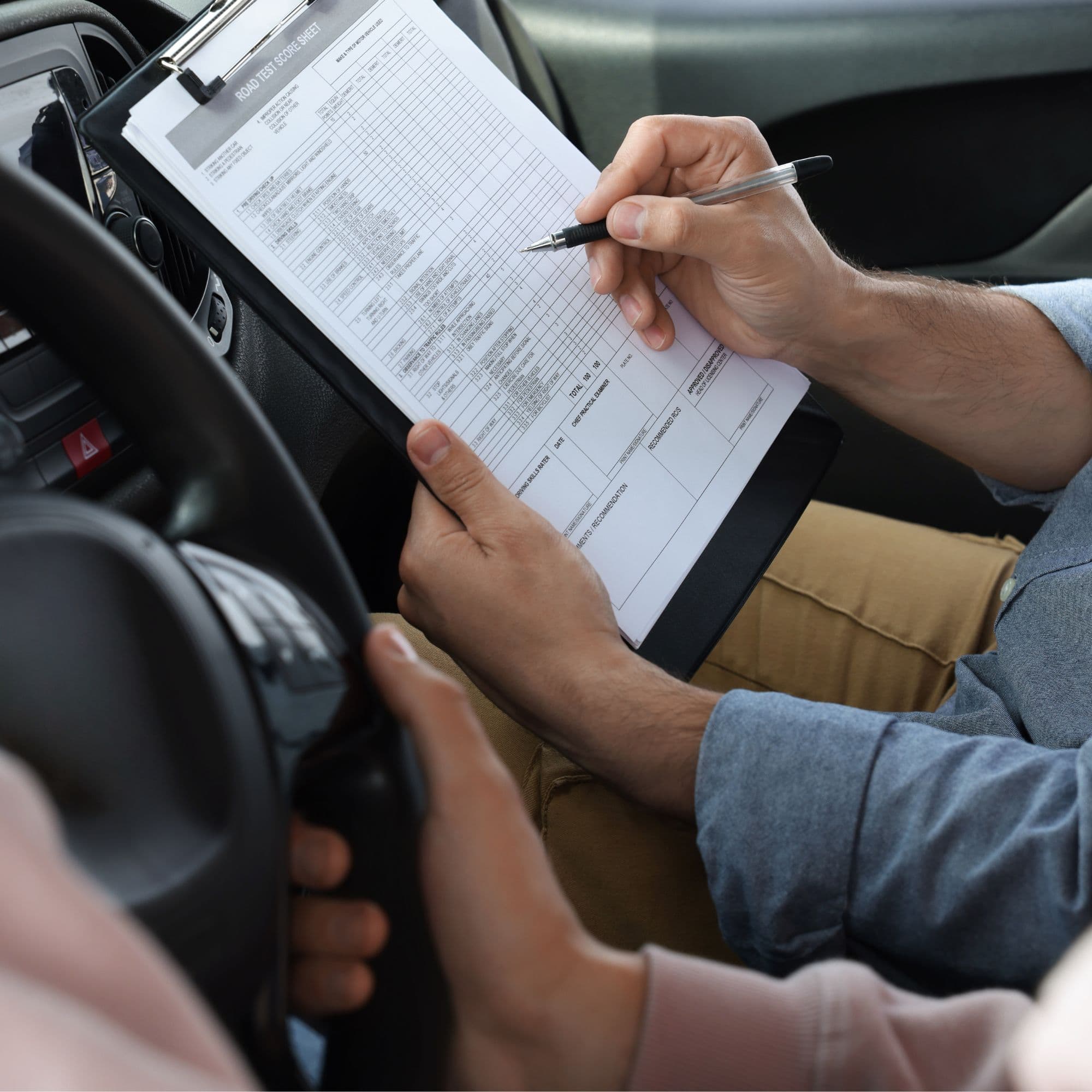 Driving examiner holds a road test score sheet and pen next to a student driver.